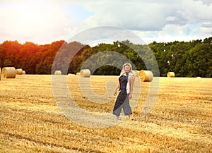 A young Caucasian blond woman with a smile walks along a mown wheat field where there are many round large sheaves of hay.