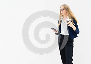 A young caucasian blonde business woman is standing in front of a white wall, looking away, and holding coffee and phone in her