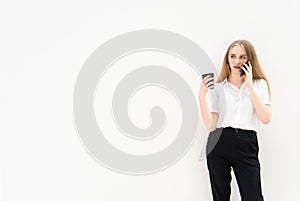 A young caucasian blonde business woman is standing in front of a white wall, talking to the phone, looking away, thinking and