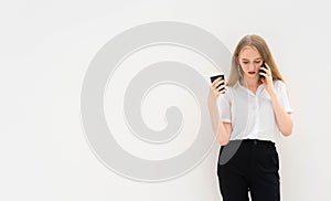 A young caucasian blonde business woman is standing in front of a white wall, talking to the phone, looking down, thinking and