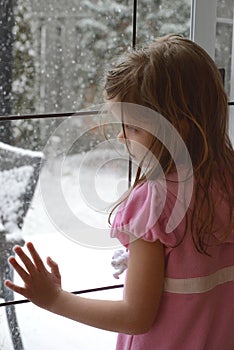 The young Caucasian blonde girl leans on the french glass door and looks at the falling snowflakes outside.