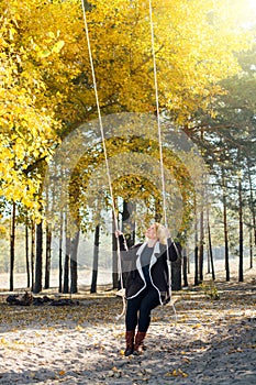 Young caucasian blonde woman in brown cardigan riding a swing in yellow autumn forest. Vertical orientation