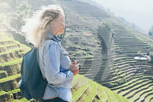 Young Caucasian blonde woman in denim shirt taking pictures of Sapa rice terraces at sunset in Lao Cai province, Vietnam