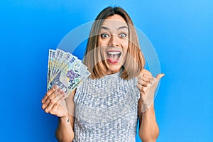 Young caucasian blonde woman holding 100 romanian leu banknotes pointing thumb up to the side smiling happy with open mouth