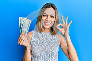 Young caucasian blonde woman holding bangladesh taka banknotes doing ok sign with fingers, smiling friendly gesturing excellent