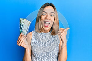 Young caucasian blonde woman holding bangladesh taka banknotes pointing thumb up to the side smiling happy with open mouth