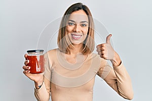 Young caucasian blonde woman holding jam smiling happy and positive, thumb up doing excellent and approval sign