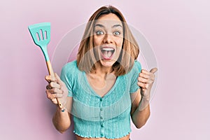 Young caucasian blonde woman holding silicone spatula pointing thumb up to the side smiling happy with open mouth
