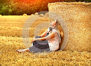 Young caucasian blonde woman sitting on a mown wheat field near a huge sheaf of hay enjoying nature at sunset. People and travel.