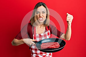 Young caucasian blonde woman wearing cook apron cooking meat on pan annoyed and frustrated shouting with anger, yelling crazy with