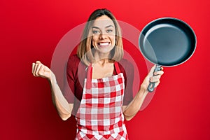 Young caucasian blonde woman wearing cook apron holding pan screaming proud, celebrating victory and success very excited with