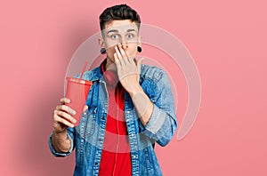 Young caucasian boy with ears dilation drinking glass of cola beverage covering mouth with hand, shocked and afraid for mistake