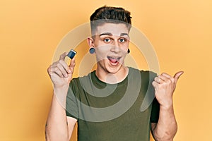 Young caucasian boy with ears dilation holding sdxc card pointing thumb up to the side smiling happy with open mouth