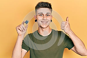 Young caucasian boy with ears dilation holding sdxc card smiling happy and positive, thumb up doing excellent and approval sign