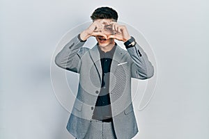 Young caucasian boy with ears dilation wearing business jacket doing heart shape with hand and fingers smiling looking through