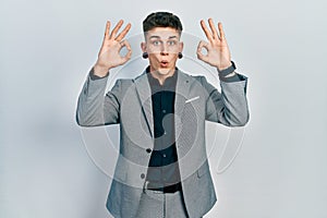 Young caucasian boy with ears dilation wearing business jacket looking surprised and shocked doing ok approval symbol with fingers