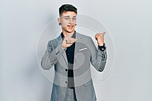 Young caucasian boy with ears dilation wearing business jacket pointing to the back behind with hand and thumbs up, smiling