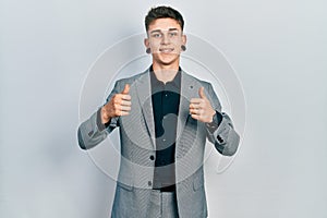 Young caucasian boy with ears dilation wearing business jacket success sign doing positive gesture with hand, thumbs up smiling