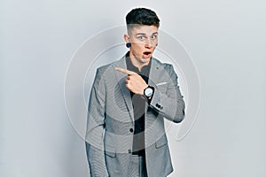 Young caucasian boy with ears dilation wearing business jacket surprised pointing with finger to the side, open mouth amazed