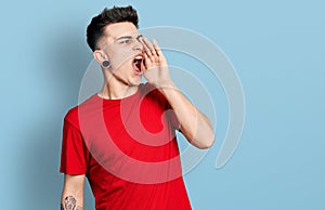 Young caucasian boy with ears dilation wearing casual red t shirt shouting and screaming loud to side with hand on mouth