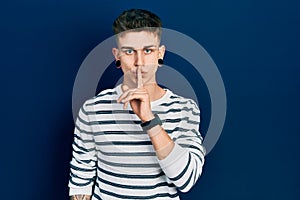 Young caucasian boy with ears dilation wearing casual striped shirt asking to be quiet with finger on lips