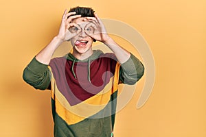 Young caucasian boy with ears dilation wearing casual sweatshirt doing ok gesture like binoculars sticking tongue out, eyes