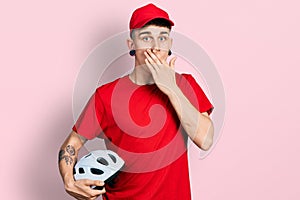 Young caucasian boy with ears dilation wearing delivery uniform and cap holding bike helmet covering mouth with hand, shocked and