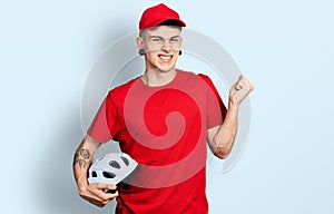 Young caucasian boy with ears dilation wearing delivery uniform and cap holding bike helmet screaming proud, celebrating victory