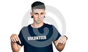 Young caucasian boy with ears dilation wearing volunteer t shirt pointing down looking sad and upset, indicating direction with