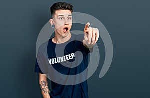 Young caucasian boy with ears dilation wearing volunteer t shirt pointing with finger surprised ahead, open mouth amazed