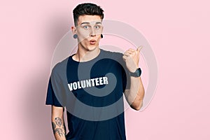 Young caucasian boy with ears dilation wearing volunteer t shirt surprised pointing with hand finger to the side, open mouth