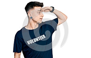 Young caucasian boy with ears dilation wearing volunteer t shirt very happy and smiling looking far away with hand over head