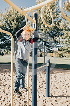 Young Caucasian boy hanging on monkey bars in park on playground. Summer outdoors activity for kids. Active preschool child doing