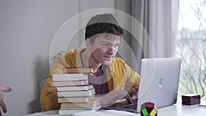Young Caucasian brunette boy playing on laptop as his mother coming up with stack of books. Woman putting literature on