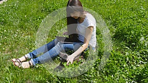 Young caucasian brunette girl sitting on grass on the meadow in the park, stroking a cat and using digital tablet pc.