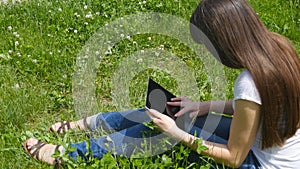 Young caucasian brunette girl sitting on grass on the meadow in the park and using digital tablet pc. Happy woman