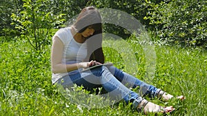 Young caucasian brunette girl sitting on grass on the meadow in the park and using digital tablet pc. Happy woman