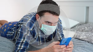 Young Caucasian brunette man wear turquoise disposable medical protective face mask, plaid shirt, blue panties lies on sofa