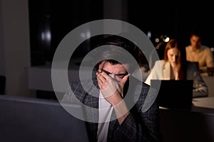 young caucasian brunnete man in suit sits at night in office, eyes hurt from overexertion