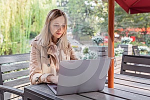 Young Caucasian business woman with blonde hair working on laptop in outdoor cafe. College student using technology