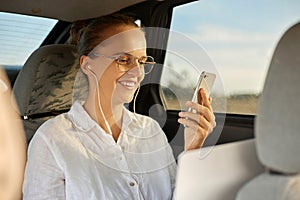 Young caucasian business woman using smart phone and smiling while sitting on back seat in the car browsing internet reading