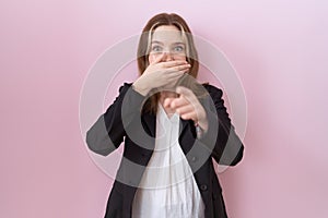 Young caucasian business woman wearing black jacket laughing at you, pointing finger to the camera with hand over mouth, shame