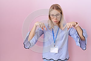 Young caucasian business woman wearing id card pointing down looking sad and upset, indicating direction with fingers, unhappy and