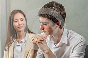 Young caucasian businessman looks seriously working, beautiful woman relax looking at man, sitting, selective focus at businessman