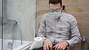 Young Caucasian businessman wearing plaid gray shirt, black glasses, protective medical face mask, sit on toilet bowl