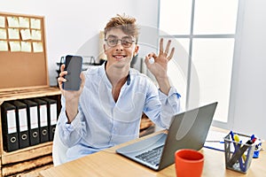 Young caucasian businessman working using laptop and showing smartphone doing ok sign with fingers, smiling friendly gesturing