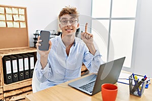 Young caucasian businessman working using laptop and showing smartphone smiling with an idea or question pointing finger with