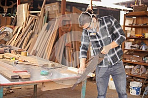 Young Caucasian carpenter man is sawing plank of wood in his own garage style workshop for hobby