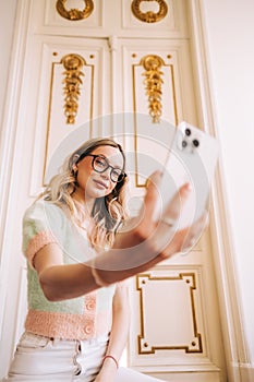 Young caucasian cheerful woman using mobile phone in bright living room, connecting to friends in video call or taking selfie