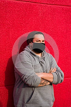 Young caucasian chubby man with a hat and face mask posing against a red wall with arms crossed